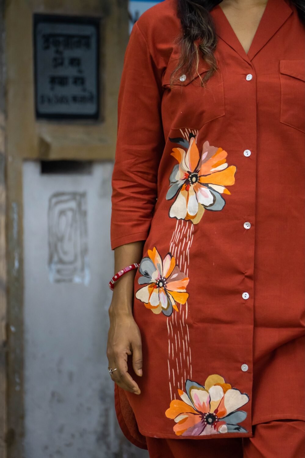 lose-up of three large artisan hand-painted floral motifs in ochre, cream, grey, and orange with vertical white line detailing on a rust orange button-down blazer tunic