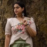 Model posing against a tree trunk in a beige hand-painted floral scallop top with curved hem and sage green trousers.