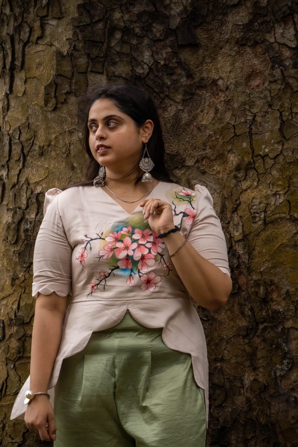 Model posing against a tree trunk in a beige hand-painted floral scallop top with curved hem and sage green trousers.