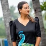 Portrait view of a woman wearing a black V-neck Malai Cotton dress with hand painted blue peacock motif