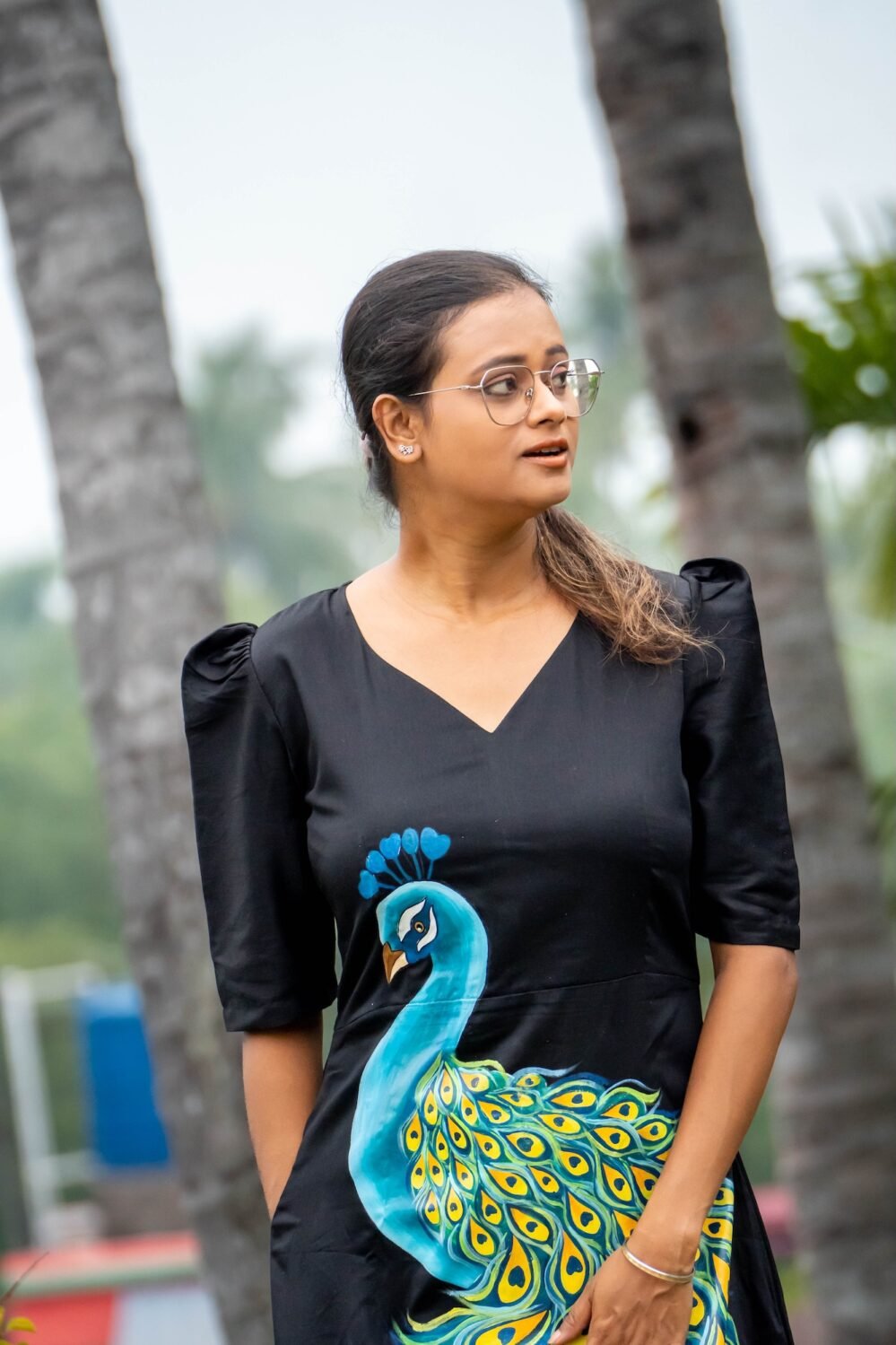 Portrait view of a woman wearing a black V-neck Malai Cotton dress with hand painted blue peacock motif
