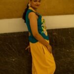 Side view of a woman wearing a teal waistcoat with hand-painted orange citrus motifs and bright yellow harem pants leaning against a textured wall