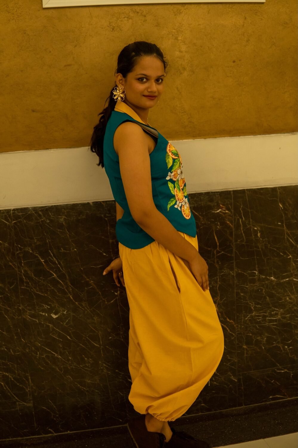 Side view of a woman wearing a teal waistcoat with hand-painted orange citrus motifs and bright yellow harem pants leaning against a textured wall