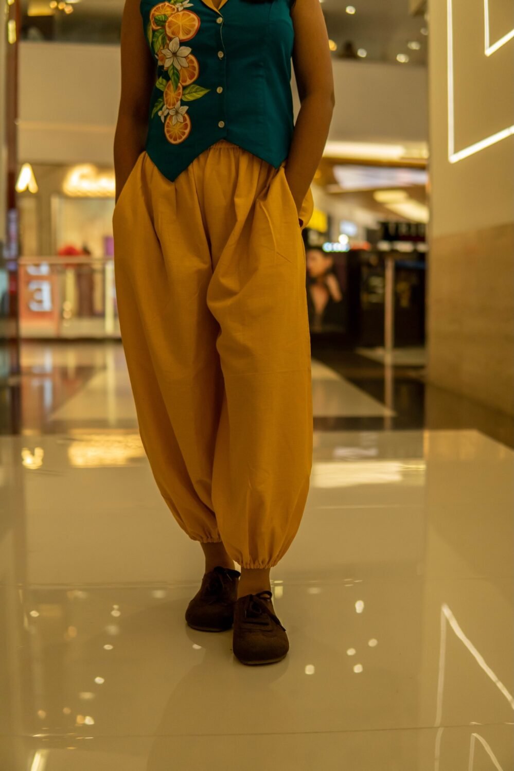 Close-up of gathered yellow harem pants with an elasticated ankle cuff, paired with brown casual shoes on a reflective indoor floor