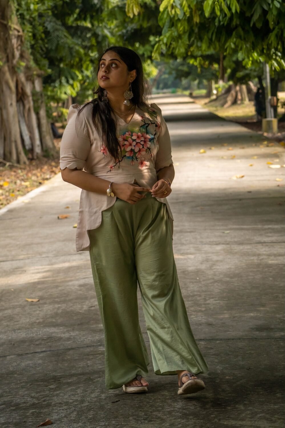 Candid full-length shot of a woman looking away, wearing a hand-painted beige blossom scallop top and sage green trousers.