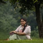 Woman sitting on green park grass showcasing a beige hand-painted blossom scallop top and sage green trouser co-ord set.