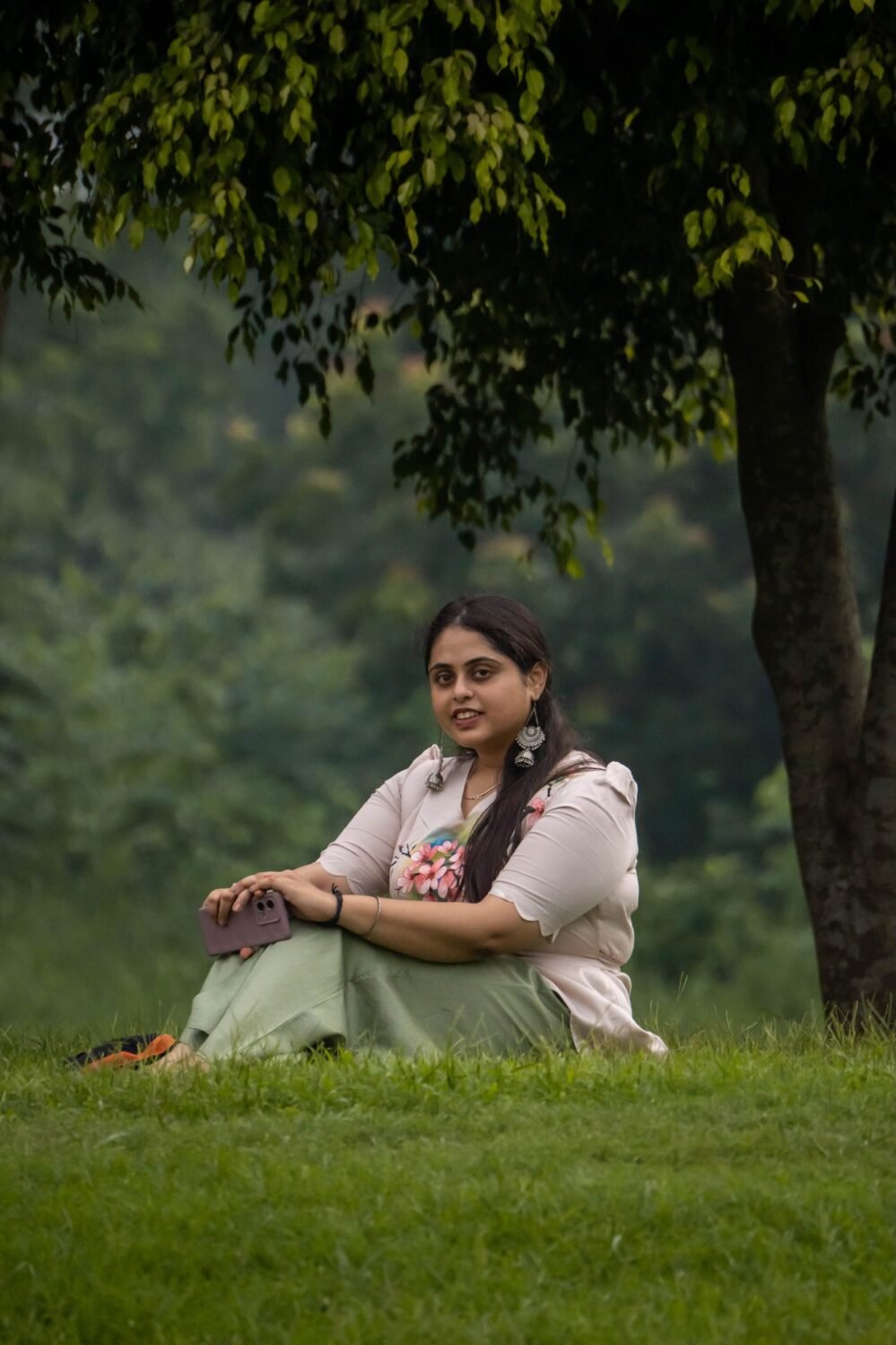 Woman sitting on green park grass showcasing a beige hand-painted blossom scallop top and sage green trouser co-ord set.