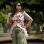 Three-quarter length shot of a woman in sunglasses posing outdoors in a beige hand-painted floral scallop top and sage green trousers.