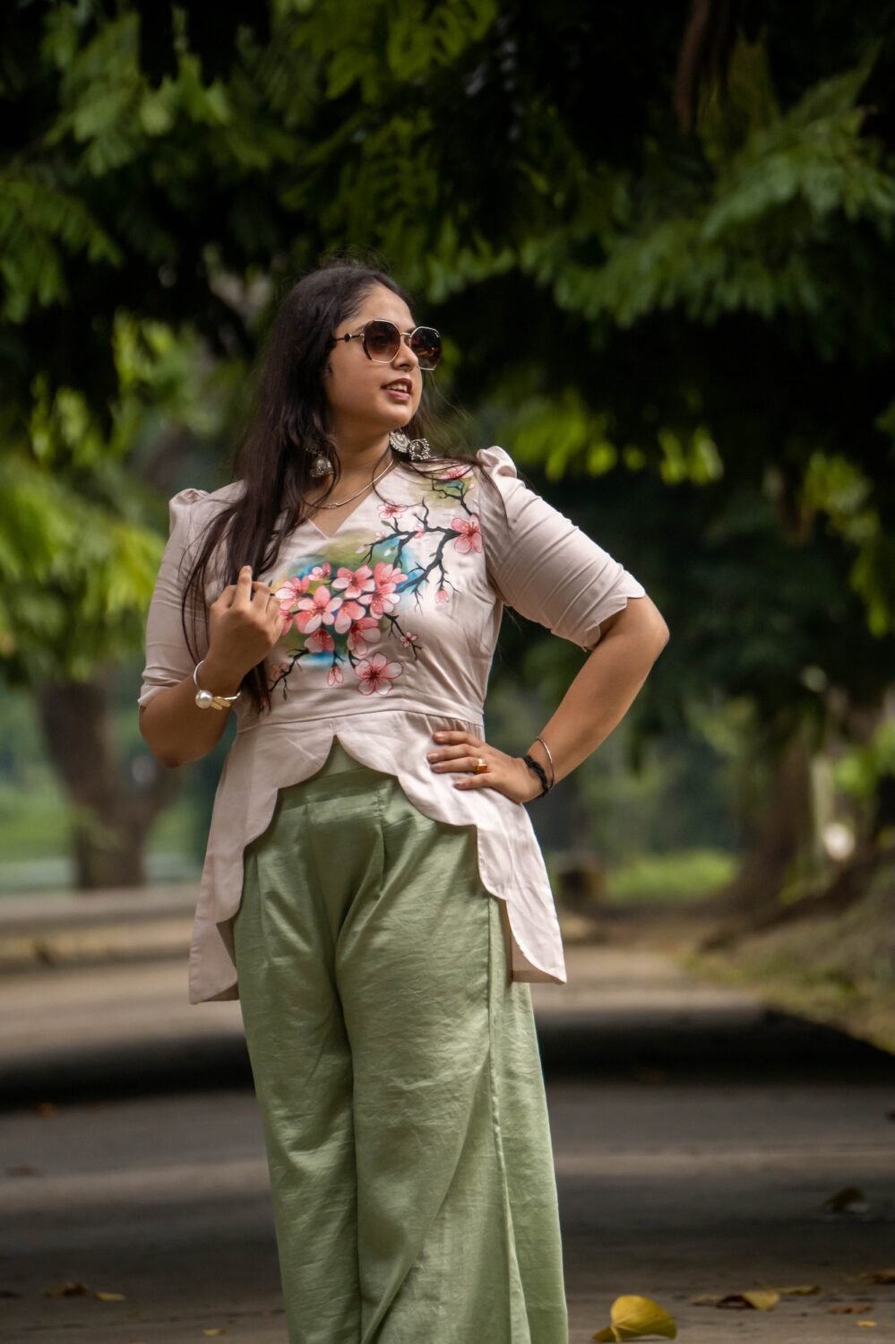 Three-quarter length shot of a woman in sunglasses posing outdoors in a beige hand-painted floral scallop top and sage green trousers.
