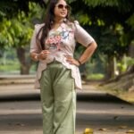 Full-length outdoor shot of a woman wearing a beige hand-painted cherry blossom scallop top paired with relaxed sage green wide-leg pants.