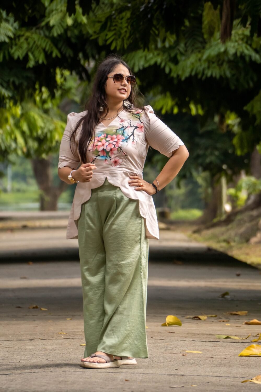 Full-length outdoor shot of a woman wearing a beige hand-painted cherry blossom scallop top paired with relaxed sage green wide-leg pants.