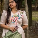 Woman leaning against a tree trunk wearing a beige hand-painted floral scallop top and sage green trousers.