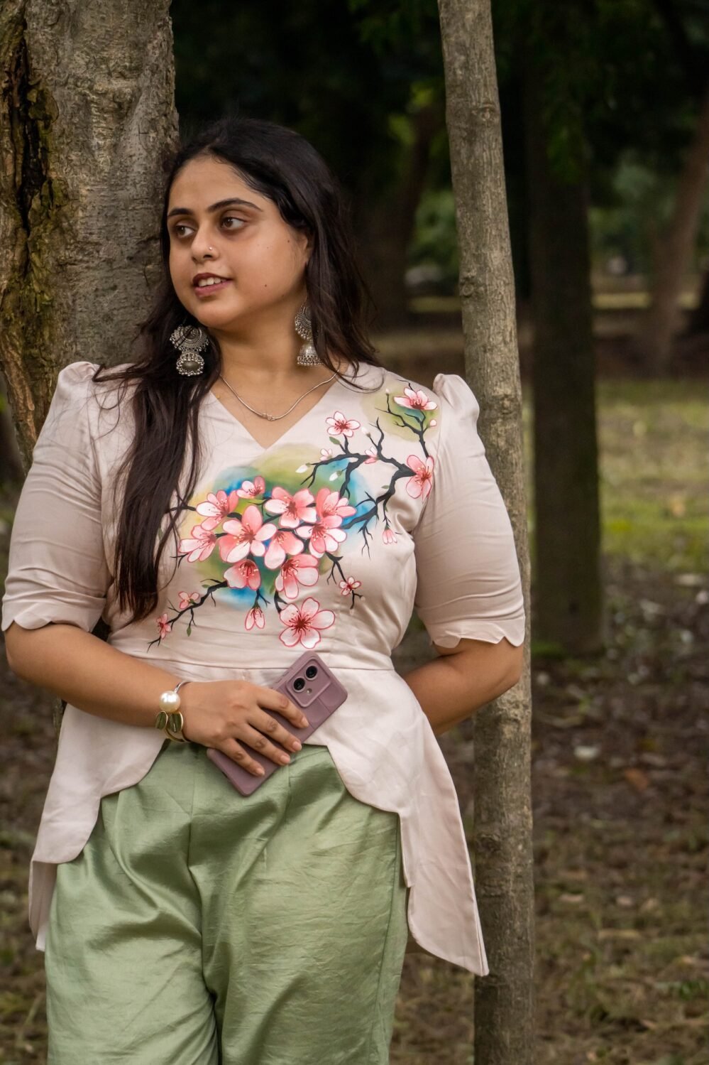 Woman leaning against a tree trunk wearing a beige hand-painted floral scallop top and sage green trousers.