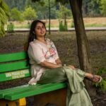 Woman seated on a garden bench showcasing a hand-painted beige blossom scallop top with sage green wide-leg trousers.