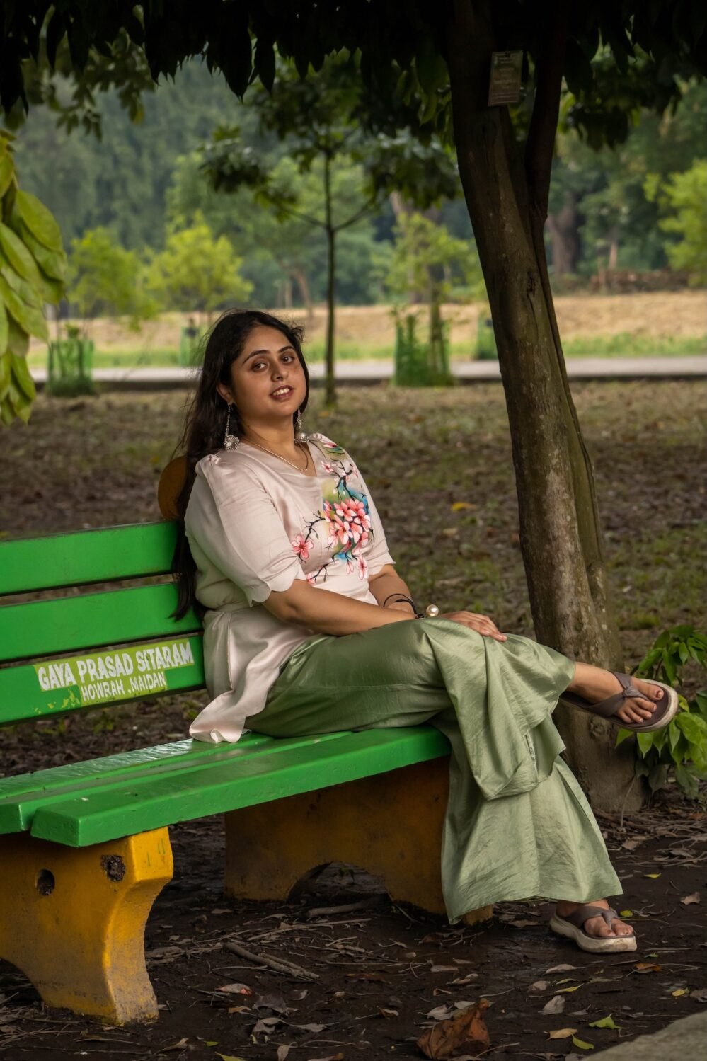 Woman seated on a garden bench showcasing a hand-painted beige blossom scallop top with sage green wide-leg trousers.