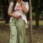 Full-length view of a woman leaning against a tree trunk wearing a beige hand-painted floral scallop top and sage green flowy trousers.