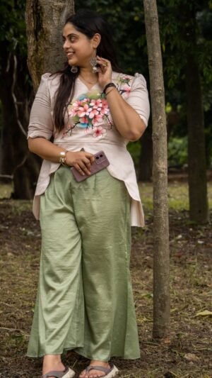 Full-length view of a woman leaning against a tree trunk wearing a beige hand-painted floral scallop top and sage green flowy trousers.