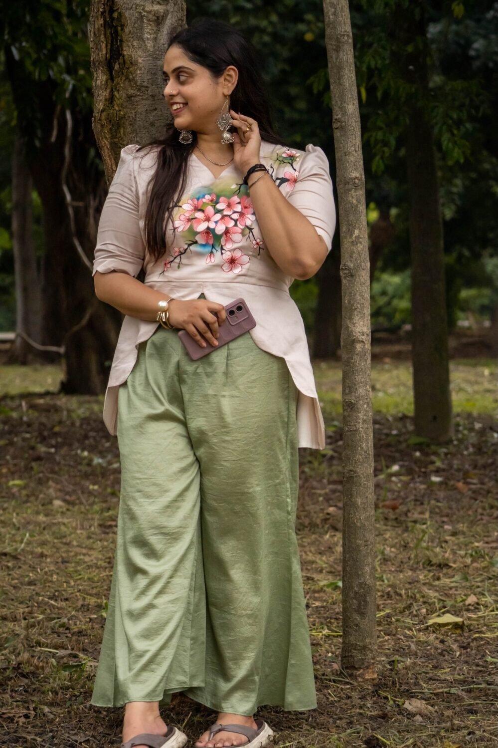Full-length view of a woman leaning against a tree trunk wearing a beige hand-painted floral scallop top and sage green flowy trousers.