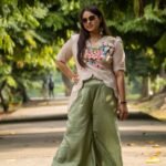 Full-length pose of a woman wearing a beige hand-painted floral scallop top and wide-leg sage green trousers in a park setting.