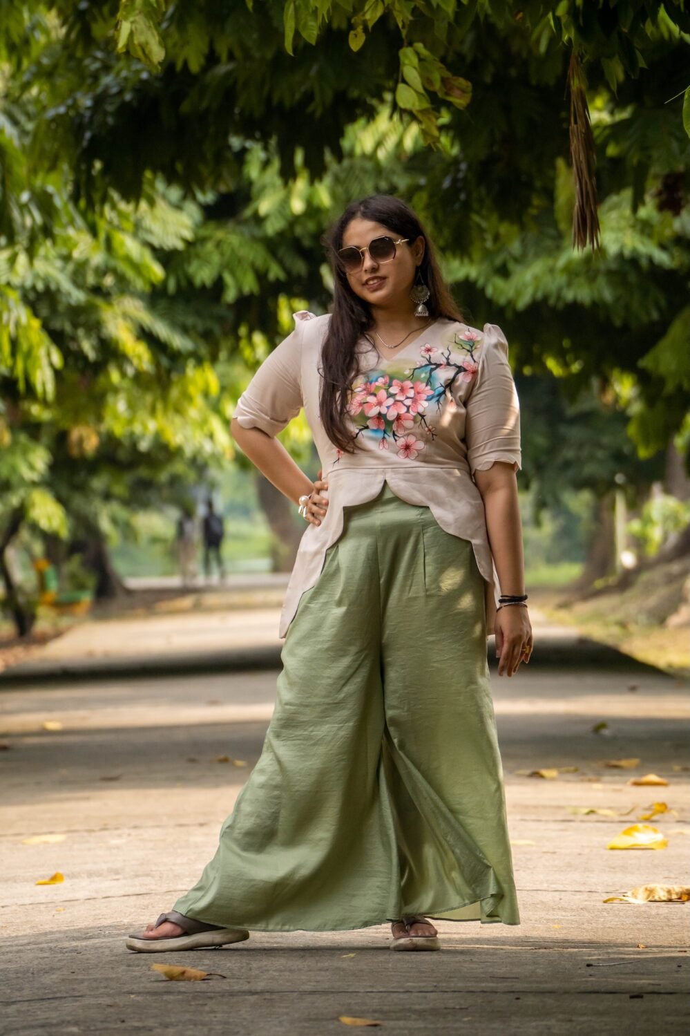 Full-length pose of a woman wearing a beige hand-painted floral scallop top and wide-leg sage green trousers in a park setting.