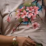 Close-up detail shot of custom hand-painted pink blossom floral artwork on a beige scallop top with a pearl bracelet accessory.