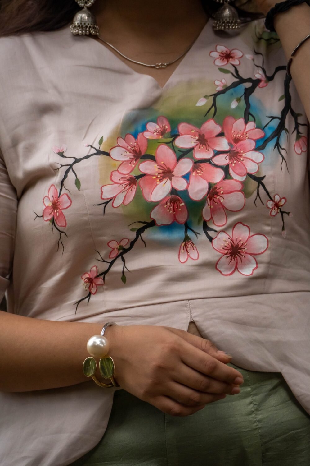 Close-up detail shot of custom hand-painted pink blossom floral artwork on a beige scallop top with a pearl bracelet accessory.