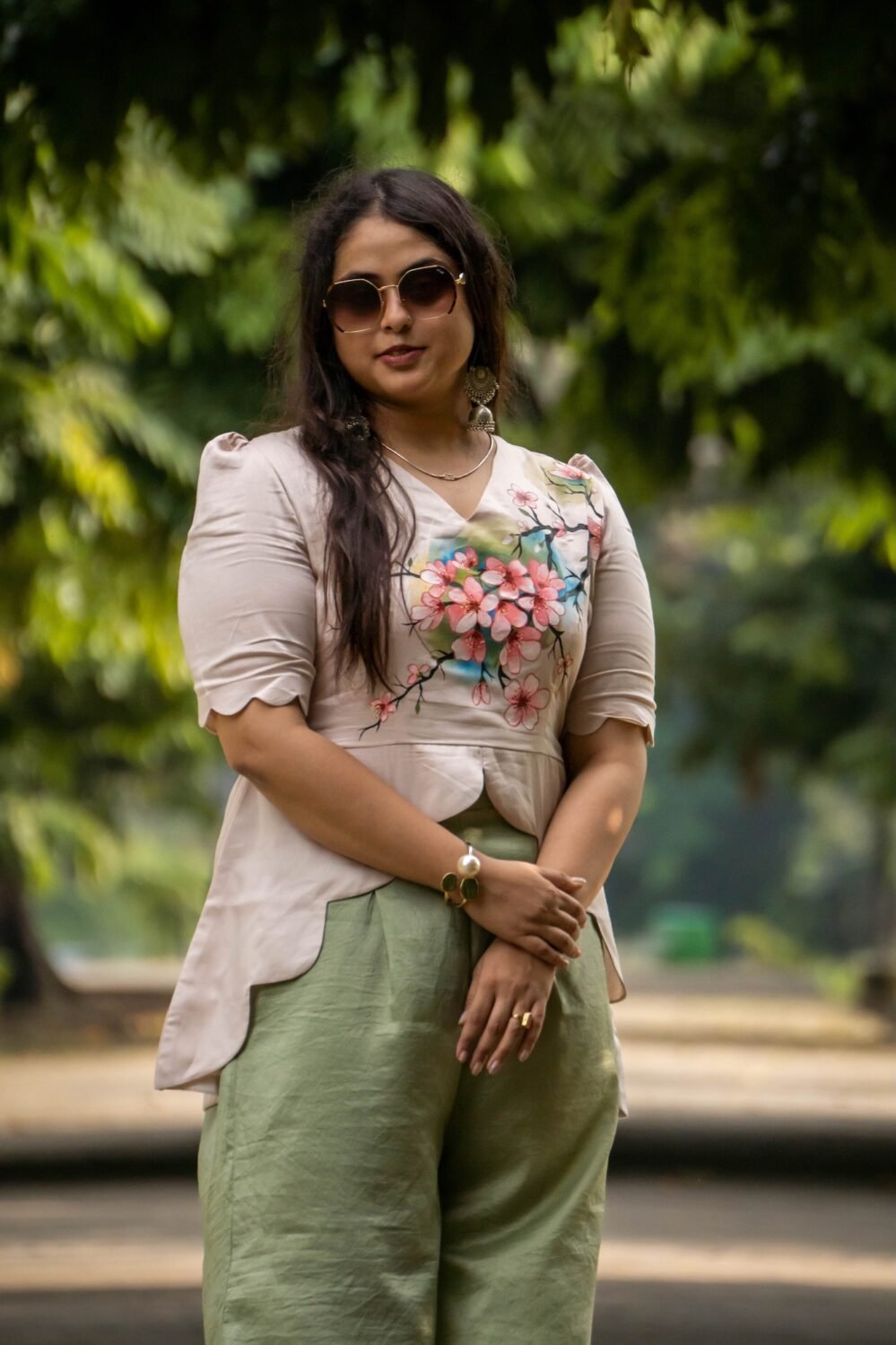 Medium close-up of a woman in sunglasses showing the detailed hand-painted floral art across her beige scallop top with scalloped sleeves.