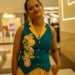 A woman smiling indoors wearing a sleeveless teal waistcoat featuring a vibrant hand-painted floral and orange slice design down the front