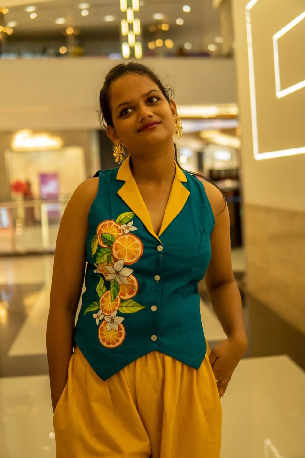 A woman smiling indoors wearing a sleeveless teal waistcoat featuring a vibrant hand-painted floral and orange slice design down the front