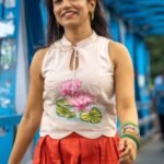 Smiling model walking on a blue bridge wearing an off-white hand-painted lotus top and red pleated skirt