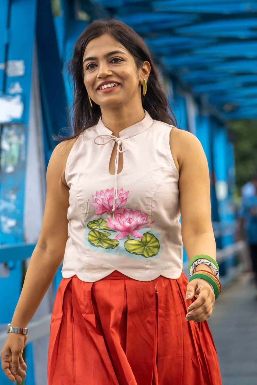 Smiling model walking on a blue bridge wearing an off-white hand-painted lotus top and red pleated skirt