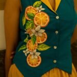 Detailed close-up of a teal waistcoat showcasing intricate hand-painted orange slices, green leaves, and white blossoms along the button placket