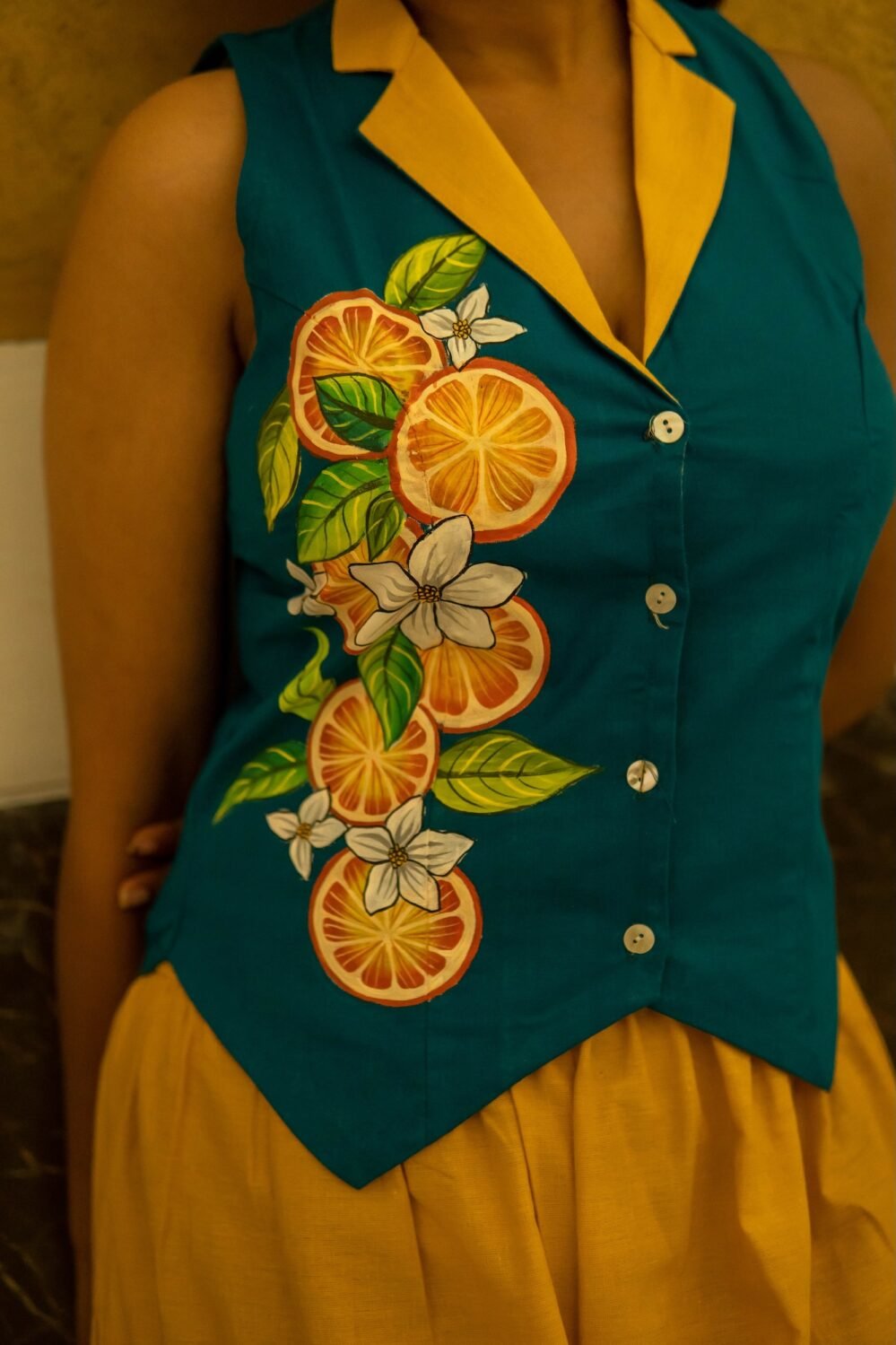 Detailed close-up of a teal waistcoat showcasing intricate hand-painted orange slices, green leaves, and white blossoms along the button placket