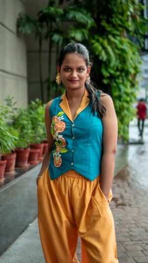 Front view of a woman posing outdoors in a teal vest with hand-painted citrus details, a prominent yellow collar, and relaxed yellow trousers