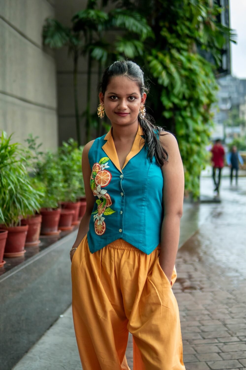 Front view of a woman posing outdoors in a teal vest with hand-painted citrus details, a prominent yellow collar, and relaxed yellow trousers