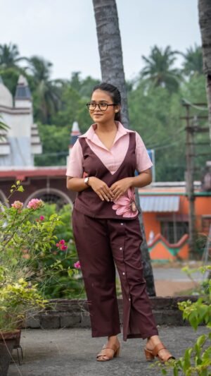 A woman wearing the Kshiti hand-painted cotton co-ord set. The outfit includes a short-sleeve chocolate brown top with a peach collar and a detailed, hand-painted pink floral motif on the side, paired with matching brown cotton trousers featuring decorative white buttons along the overlap front crease.