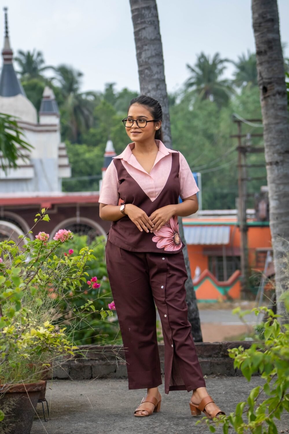 A woman wearing the Kshiti hand-painted cotton co-ord set. The outfit includes a short-sleeve chocolate brown top with a peach collar and a detailed, hand-painted pink floral motif on the side, paired with matching brown cotton trousers featuring decorative white buttons along the overlap front crease.