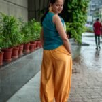 Back view of a woman walking outdoors wearing a teal sleeveless top and loosely fitted yellow pants next to a row of potted plants