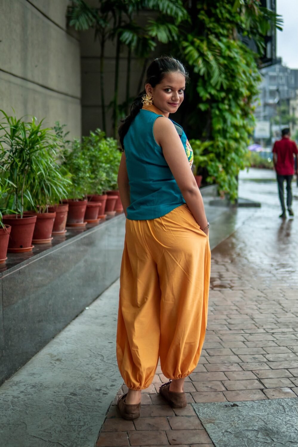 Back view of a woman walking outdoors wearing a teal sleeveless top and loosely fitted yellow pants next to a row of potted plants