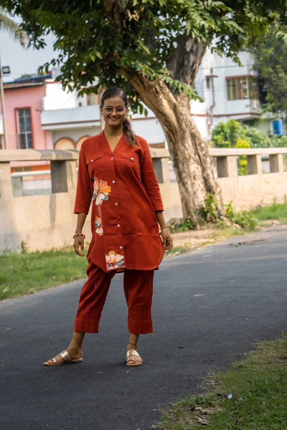 Full shot of a model standing on an outdoor road in a premium terracotta cotton co-ord set, featuring a structured dual-pocket longline blazer top and pleated hem crop trousers.