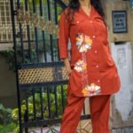 Front view of a model standing by a black iron gate posing in a rust orange fusion co-ord set with hand-painted floral designs on the long button-down top and matching cropped trousers