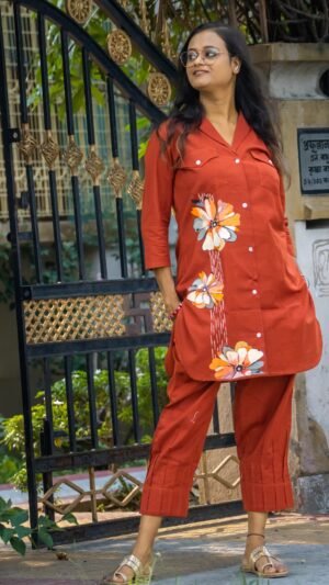 Front view of a model standing by a black iron gate posing in a rust orange fusion co-ord set with hand-painted floral designs on the long button-down top and matching cropped trousers