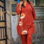 Full-length view of a model walking outdoors in a rust orange two-piece co-ord set featuring a longline hand-painted floral blazer top and straight-leg trousers.