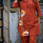 A model wearing a hand-painted rust orange longline shirt blazer tunic and matching trousers walking near a textured wall, showcasing the modern silhouette and comfortable fit