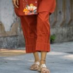 Close-up shot focused on the lower portion of terracotta trouser pants, displaying the clean structural pintuck pleat lines and geometric hem design above gold flat sandals.