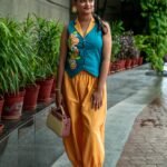 A woman carrying a small handbag and posing outdoors in a teal waistcoat featuring hand-painted citrus fruits and matching yellow harem pants