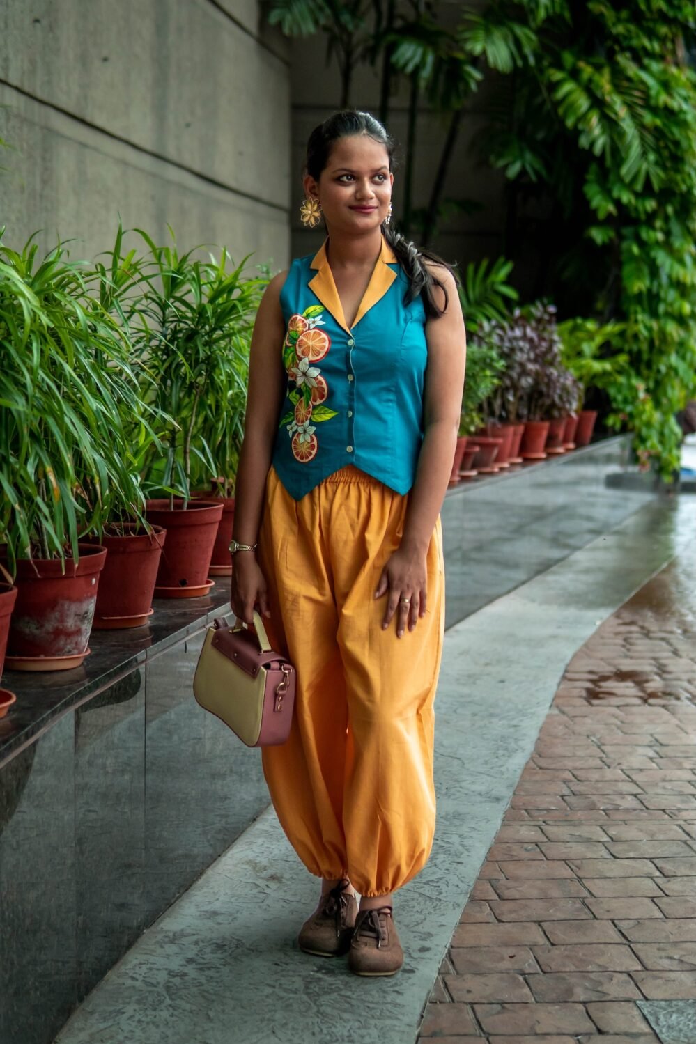 A woman carrying a small handbag and posing outdoors in a teal waistcoat featuring hand-painted citrus fruits and matching yellow harem pants