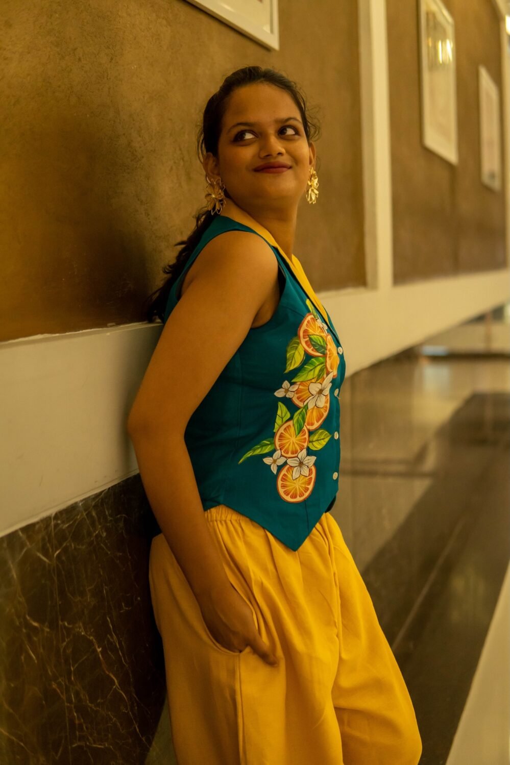Side angle shot of a woman looking over her shoulder indoors, highlighting the hand-painted orange fruit pattern on her teal waistcoat