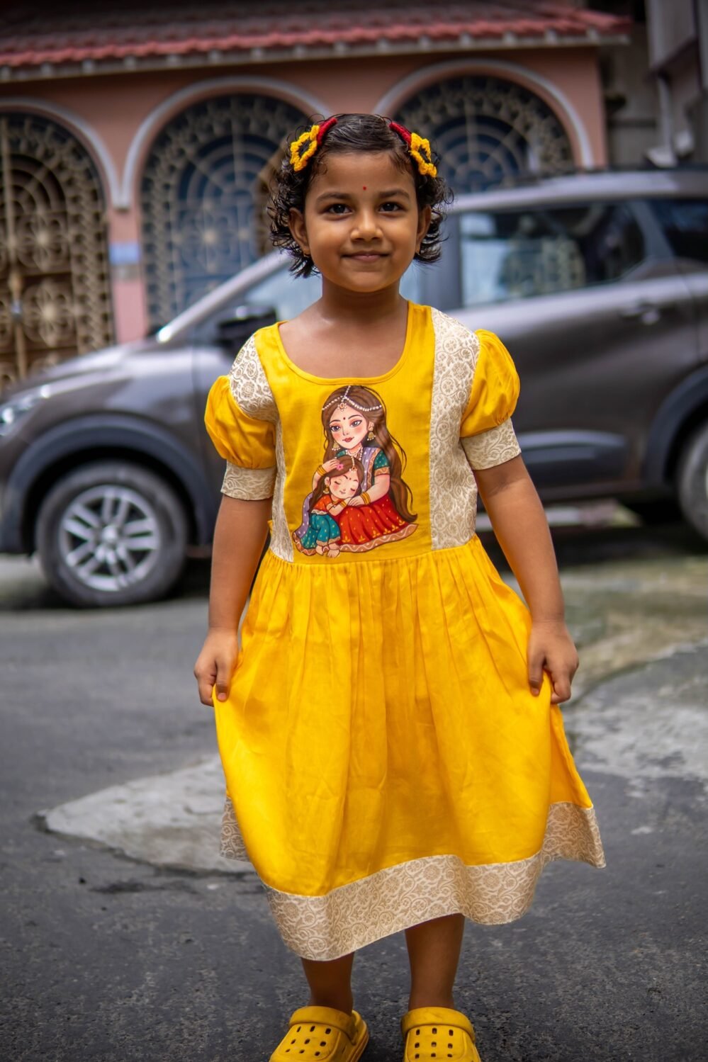 Front view of a child standing outdoors in the yellow handpainted Durga Dulali cotton frock featuring brocade accents.