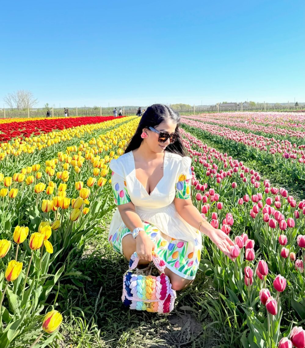 Woman crouching in a yellow tulip field touching pink tulips while wearing a handpainted floral short dress and sunglasses